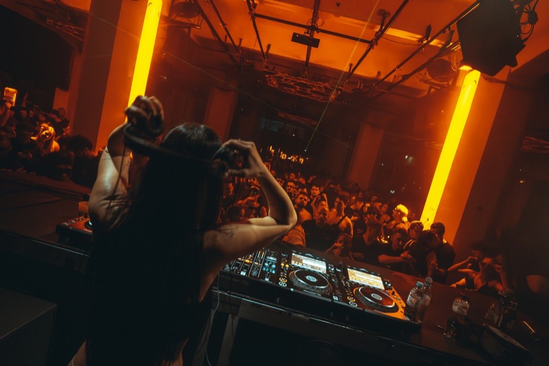 DJ with arms raised behind the decks, crowd in orange-lit room.