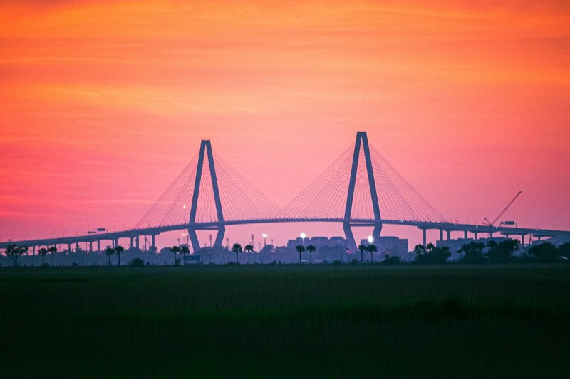 The Arthur Ravenel Jr. Bridge silhouetted against an orange-pink dusk sky over marsh.
