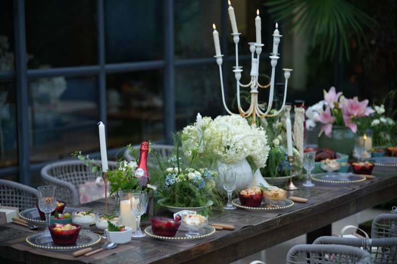 A long dinner table dressed with a candelabra, white hydrangeas, glassware and small plates at dusk.