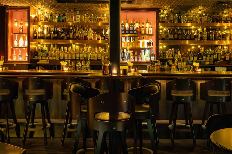 A dimly lit cocktail bar with amber back-lit bottle shelves, candles and leather stools.