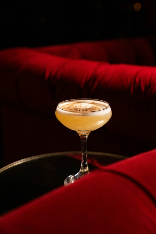 A frothy cocktail in a coupe glass on a glass table beside red velvet lounge seating.