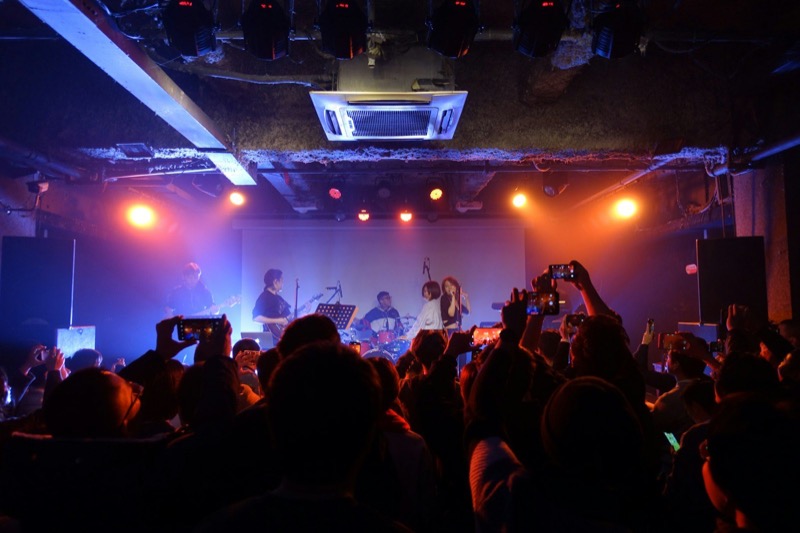 A band on a small stage in an orange-lit club, the crowd in silhouette holding up phones.